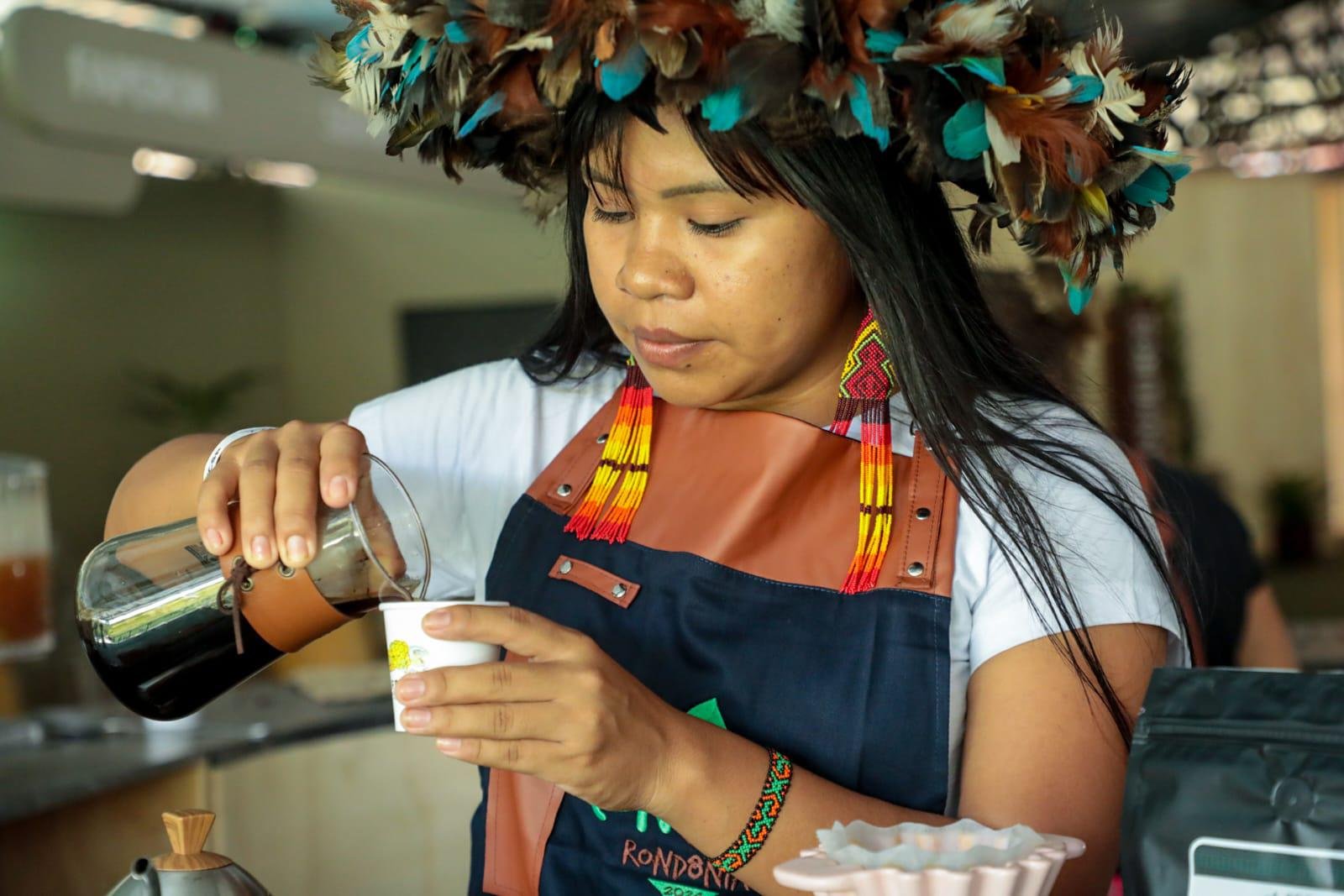 Celesty Surui prepara um café. Ela veste uma camiseta branco e um avental. Em cada orelha, tem um brinco de penas. Sobre a cabeça, um cocar cheio, também de penas